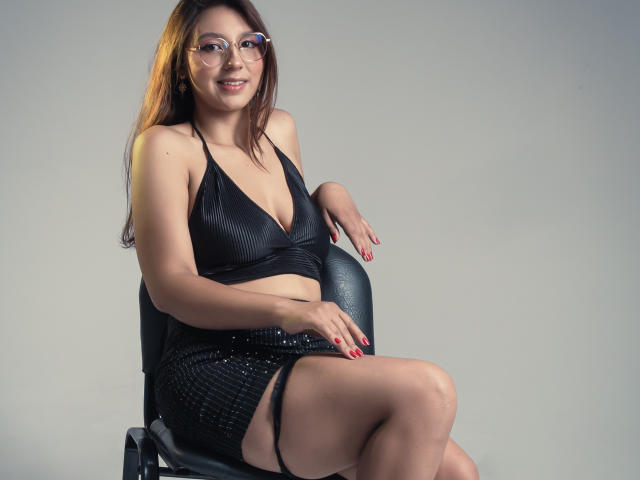 LlilthSlap, mujer asiática con gafas, top de tirantes negro, escote revelador, falda negra con lentejuelas, sentada en una silla, sonriendo, uñas rojas, piernas cruzadas, cabello castaño claro, fondo gris..