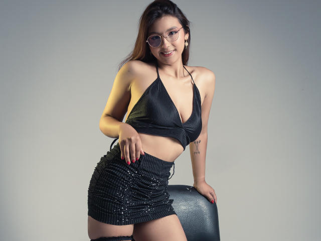 LlilthSlap with small tits, wearing a black leather halter top and a sparkly black mini-skirt, leaning on a chair, smiling at the camera, with glasses and red nails.