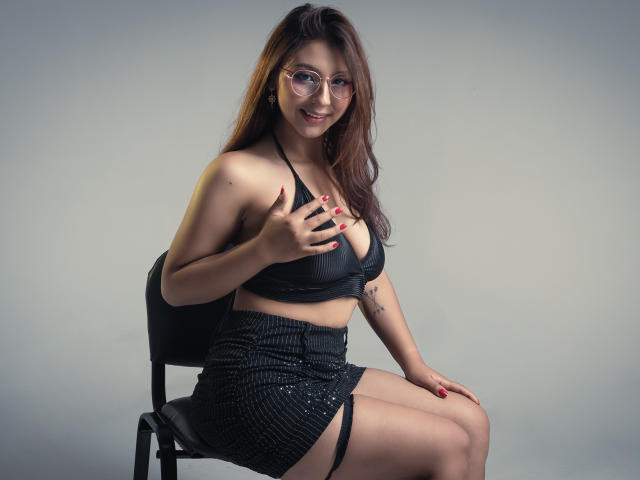 LlilthSlap con gafas, cabello castaño, sostén negro y falda a rayas, sentada en una silla, mano sobre los senos, sonriendo, tatuaje en el brazo derecho, fondo gris, iluminación de estudio..
