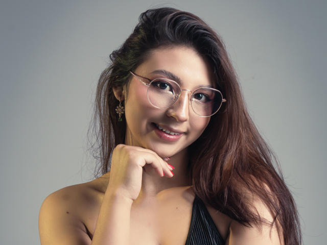 LlilthSlap has brown hair, round glasses, and a cute smile. She's touching her chin with red nails, wearing a black halter top, and has a small star earring.