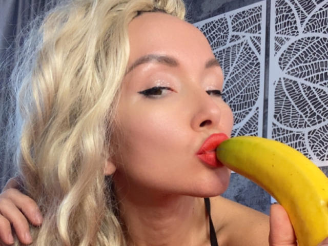 AvaGreenx with blonde hair, red lips, and black eyeliner, sucking on a yellow banana, holding it with her right hand, showing her slightly open mouth and glossy lips.