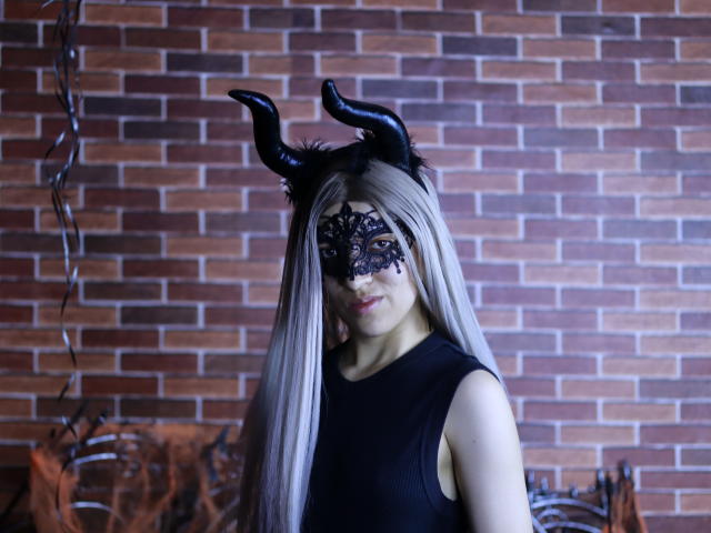 MichaelaMistress stands in front of a brick wall, wearing a black sleeveless top and a black lace mask with intricate patterns. She has long, straight silver hair and black, curved horned headwear. Her eyes are partially visible through the mask, with a neutral expression. The lighting is cool blue, highlighting her pale skin and dark outfit. The background features a metal structure on the left. The image has a cosplay or fantasy theme.