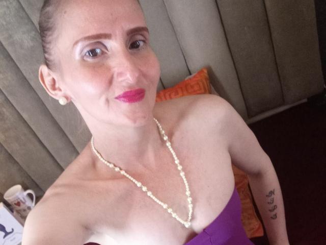 RubiAnderson with a pearl necklace, pink lipstick, and purple strapless top, showing her bare shoulders and small tits, with a tattoo on her right arm.