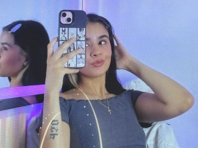 IsabellaMarynn taking a mirror selfie with a pouty face, blue off-shoulder top, cross necklace, and black hair, holding a phone with a patterned case. Her reflection is visible.