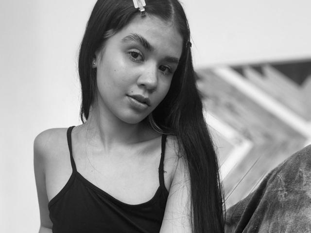Schwarz-weißes Foto von IsabellaMarynn mit langen, dunklen Haaren, die ein schwarzes Tanktop trägt und leicht zur Seite schaut, mit einem subtilen, leicht schmollenden Ausdruck. Ihre Brüste sind klein..