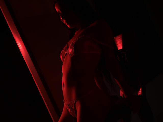 MafeSebastian è in piedi, illuminata da un semaforo rosso, indossando un reggiseno e mutandine di pizzo, mostrando i suoi seni e i suoi genitali, con una lampada soffusa che illumina lo sfondo. I suoi capezzoli sono leggermente visibili..