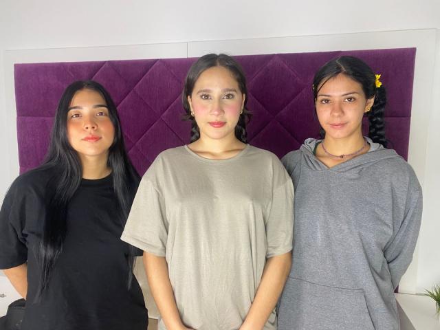Três mulheres glamourosas e atraentes, com cabelo escuro, em pé em frente a uma cabeceira roxa com padrão de diamantes. Da esquerda para a direita: camisa preta, camiseta cinza, moletom cinza. Batom vermelho em todas..