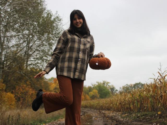 LettyCurly with long dark hair, smiling, wearing a plaid jacket, brown pants, black shoes, holding a brown stuffed toy, standing on a dirt path with autumn foliage.