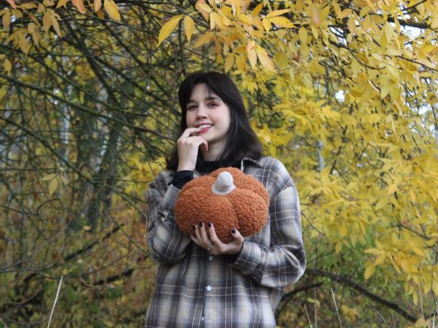 LettyCurly stands in a forest with yellow autumn leaves. She's holding a large, fuzzy brown stuffed pumpkin with a white button top. She has dark, wavy hair, light skin, and is wearing a plaid, button-up shirt. LettyCurly is smiling with her finger to her lips, looking playful. Her nails are painted dark. The texture of the stuffed pumpkin contrasts with the softness of her hair and the leafy background. The composition centers on LettyCurly, with the yellow leaves framing her upper body.