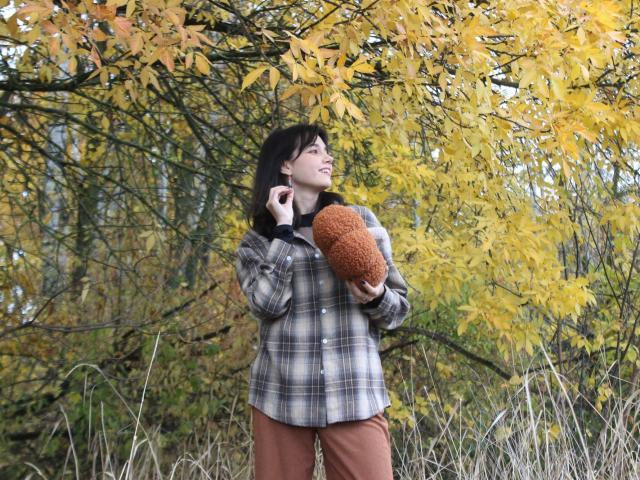 LettyCurly stands in an autumnal setting with yellow leaves. She's wearing a plaid shirt and brown pants, holding a fluffy brown pillow. Her dark hair is slightly messy, and she's looking to the side with a relaxed smile. She's holding a phone to her ear with her right hand. The image has a natural, outdoor feel with a focus on the bright yellow leaves and LettyCurly's casual, comfortable attire. The composition is balanced, with LettyCurly centered and the leafy background adding depth.