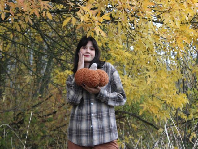 LettyCurly with brown curly hair, wearing a plaid shirt and brown pants, holding a fluffy brown stuffed animal, smiling under yellow autumn leaves.
