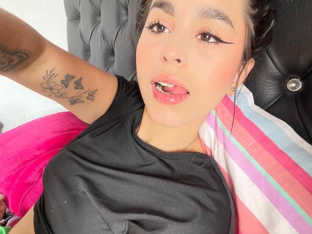 ZoeCastelli with black shirt, pink tongue out, tattoo on arm, black eyeliner, brown eyes, light skin, lying on bed with pink and striped pillow, black headboard.