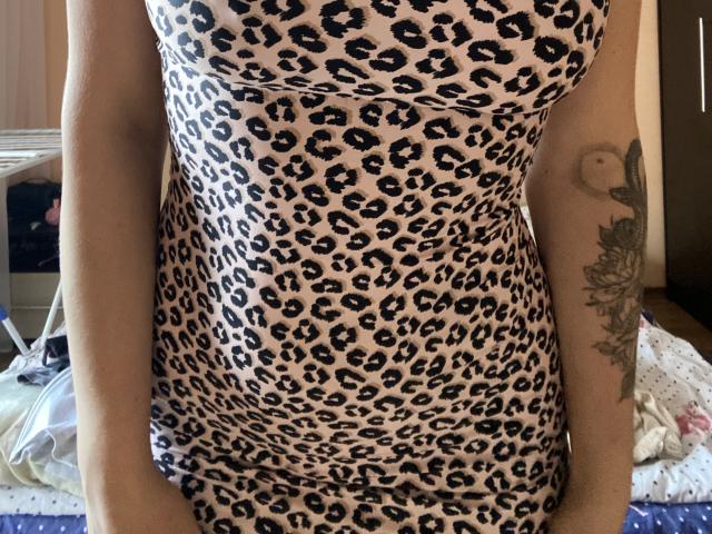 Olieri lleva un vestido ajustado de estampado de leopardo, que deja ver sus brazos con tatuajes, incluyendo un diseño floral. Se pueden ver ligeramente sus pechos a través de la tela. Está de pie en una habitación..