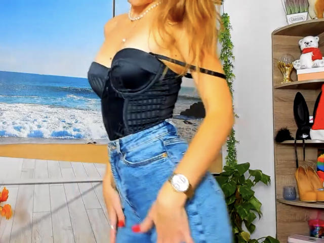 MilaYanis with blonde hair, black strapless corset, and blue high-waisted jeans, showing her tits and wearing a watch, standing in a brightly lit room.