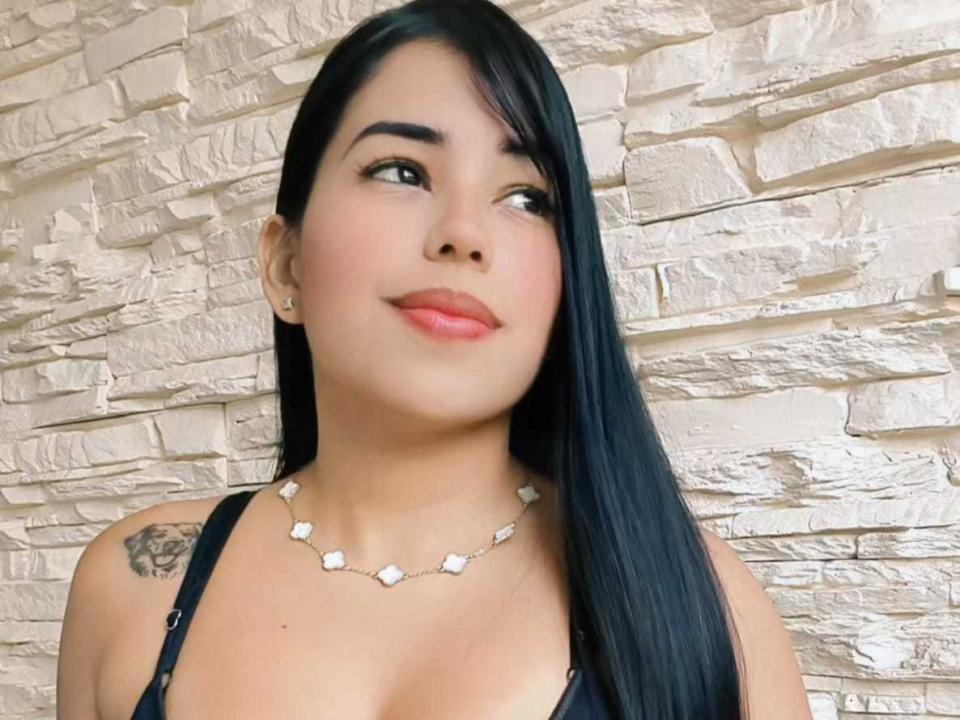 YaraGarcia has long black hair, a tattoo on her left shoulder, and a necklace with white gemstones. She's wearing a black top and looking to the side with a smile.