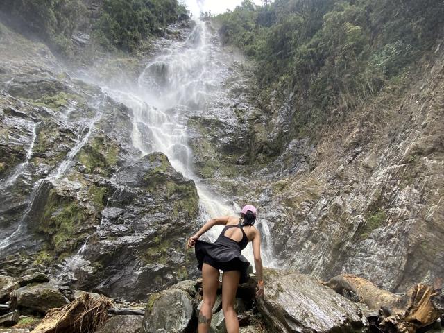 A photograph of a tall, rocky waterfall. A woman, MissValentine, stands at the bottom, looking up. She has a tan skin tone, black hair in a ponytail, and is wearing a black tank top and shorts. Her back is to the camera. The waterfall cascades down the rocky cliff, with mist at the top. The cliff is covered in greenery. She's on a large rock with smaller rocks around her. The composition is vertical.