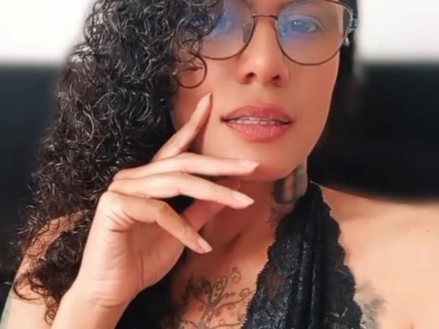 RosannaBlue with curly black hair, glasses, and pink lips, wearing a black lace top, showing a tattoo on her chest, and touching her chin.