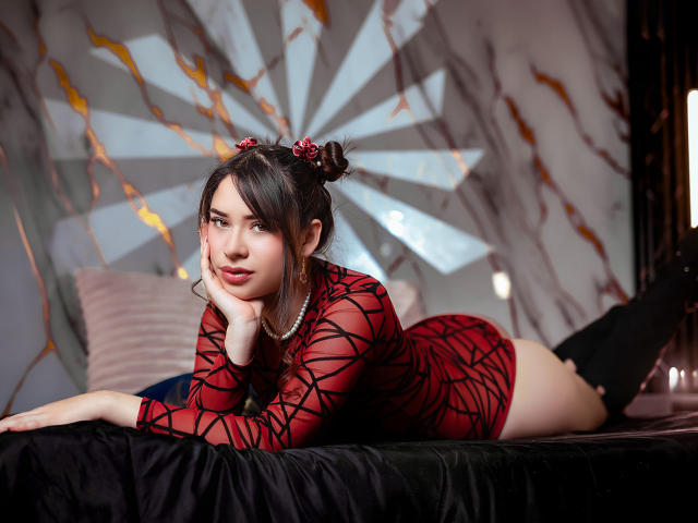 AliceOccean with dark hair in buns, wearing a red mesh dress with black lines, lying on a black bed, hand on cheek, black knee socks, pearl necklace, looking at camera.