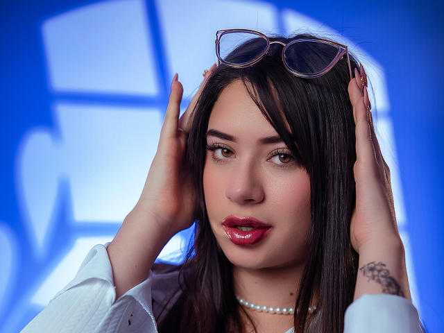 AliceOcean with hands on head, wearing glasses on forehead, white shirt, red lipstick, pearl necklace, and tattoo on right hand, against a blue background.