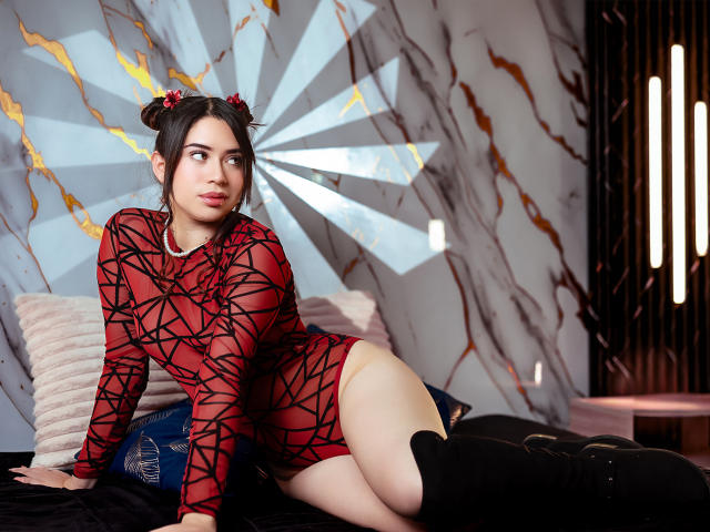 AliceOcean with dark hair in buns, red and black patterned dress, lying on bed, black thigh-highs, looking to the side, pale skin, pink lips, white pillow, blue cushion.