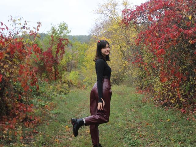 LettyCurly with dark hair, black top, and maroon pants, kicking one leg up, smiling, showing her ass, standing in a grassy path with red and yellow foliage.