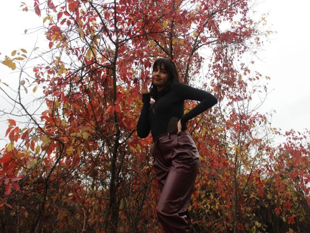 LettyCurly stands in front of a tree with vibrant red and yellow leaves. She has straight, medium-length dark hair and is wearing a black long-sleeve top and high-waisted, shiny burgundy pants. Her right hand is raised to her chin, and her left hand rests on her hip. The leafy background is dense with autumn colors. The image has a natural, slightly overcast light. LettyCurly's confident pose and colorful outfit contrast with the autumnal setting.