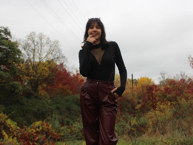 LettyCurly stands in a field with autumn foliage. She has dark hair with small red horns, wearing a black sheer long-sleeve top with a deep V-neck and maroon leather pants. Her right hand rests on her hip, left hand touches her chin, smiling. The overcast sky and trees with red, yellow, and green leaves form the background. She is centered, with a slightly relaxed posture.