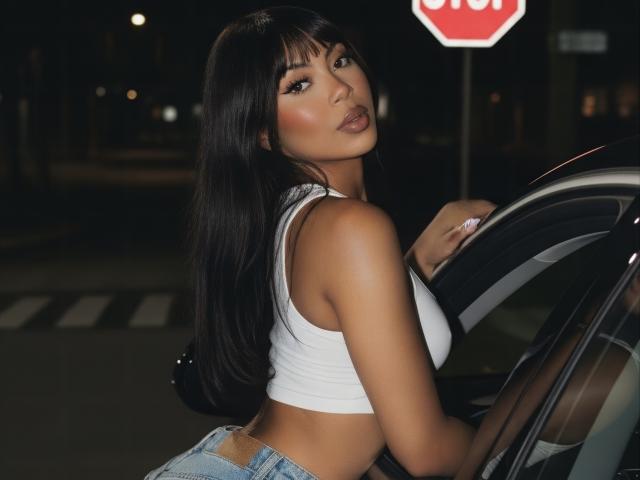 MissMaary with long black hair, wearing a white tank top and blue jeans, leaning on a car at night, showing her ass, looking back with a pouty expression.