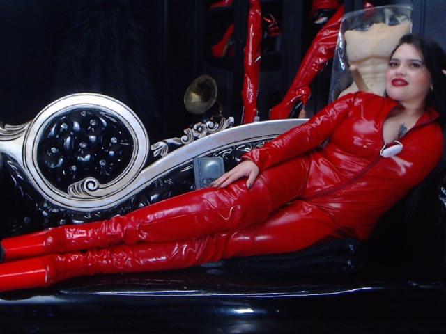 MaeveBorch in a shiny red latex bodysuit, lying on a black leather surface with her legs crossed, next to a silver and black tufted chair. Boobs visible, ass hidden.