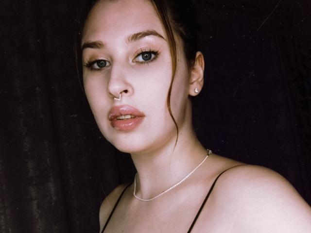 AliceLovels with nose ring, earrings, and silver necklace, looking at camera, light brown hair in a bun, wearing black spaghetti straps, slightly parted lips, pale skin, dark background.