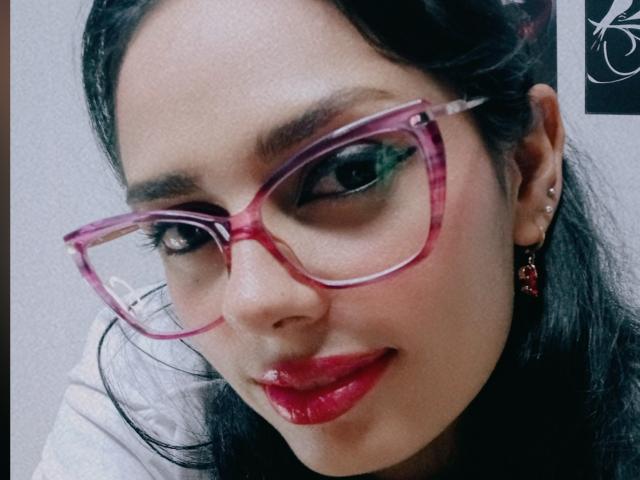 AnaisHilton with pink, transparent glasses, glossy red lips, dark hair, and a slight smile, showing a small nose piercing and red earrings, looking directly at the camera.