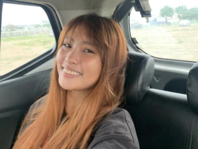 Jennelyn with long, orange-brown hair, smiling in a car, wearing a dark shirt, black leather seat behind her, looking slightly to the side, cute, small nose, white teeth.