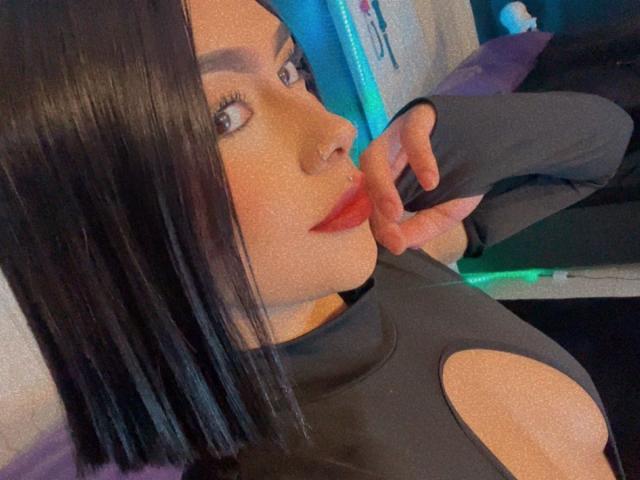 SienaReign with black shoulder-length hair, red lipstick, and nose ring, wearing a gray top with cutouts, touching her lip with a finger, looking to the side.