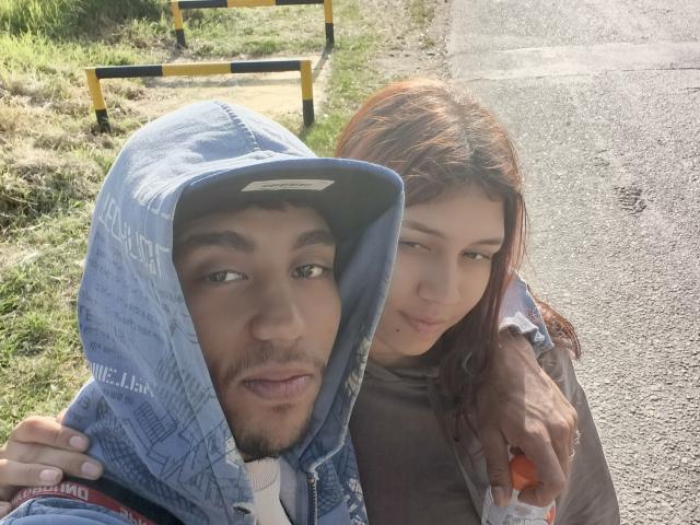 A close-up photo of AnyelinAndMark, a light-skinned woman with red hair, and a dark-skinned man with a beard, standing on a road. The man wears a blue denim hood, while AnyelinAndMark has her arm around him, holding an orange object. The man looks to the side, AnyelinAndMark looks at the camera. Bright sunlight, grassy background, yellow and black road barrier. Casual, natural, slightly blurred.