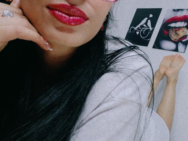 AnaisHilton with glossy red lips, black hair, wearing a white shirt, pink glasses, and a sparkling ring, lying down with her bare feet up, showing a sexy pose.