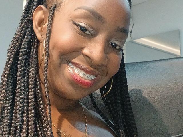 Close-up photo of KatalinaRosell. She has dark brown skin, long braided hair, and wears gold hoop earrings. Her smile reveals metallic braces on her teeth. She has red lipstick and subtle eye makeup with winged eyeliner. KatalinaRosell's hair is neatly arranged in thick braids that fall over her shoulders. She wears a gold necklace. The image has a soft, natural light that highlights her smile and facial features. Her head is slightly tilted to the right, giving a relaxed, confident vibe.