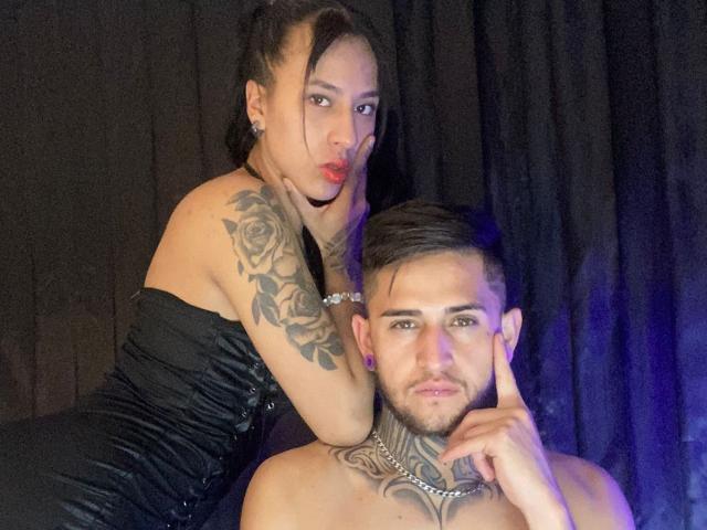 LiamAndEvelyn with rose tattoos on her arm, black dress, red lips, and Evelyn's tits pressed against Liam's neck. Liam's shirtless, tattooed, and holding Evelyn's chin.