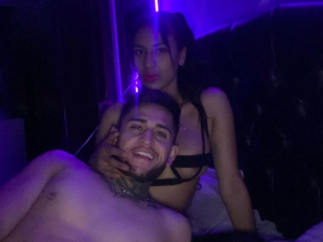 LiamAndEvelyn with tits in black bra, holding LiamAndEvelyn's head, smiling, purple light, LiamAndEvelyn's hand on LiamAndEvelyn's chest, LiamAndEvelyn's nipple visible, bed in dark room.