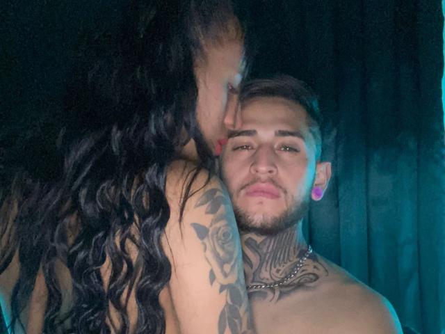 LiamAndEvelyn with long black wavy hair, kissing LiamAndEvelyn's cheek, who has tattoos, ear gauges, and a chain, with a slight smirk, both shirtless.