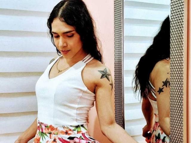 CarlyHornyDirty with long black hair, white tank top, floral skirt, star tattoo on right shoulder, standing, looking down, reflection in mirror, white wall, silver textured mirror.