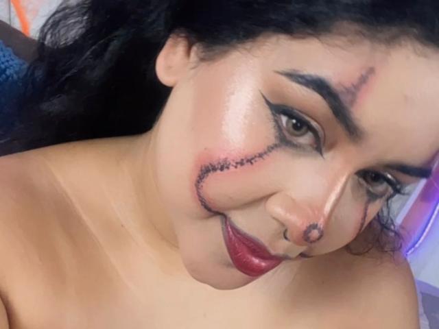 LianaAndAsaff has a stitched-up face with dark makeup, thick eyeliner, red lipstick, and a nose ring, smiling with visible scars and a puffy cheek.
