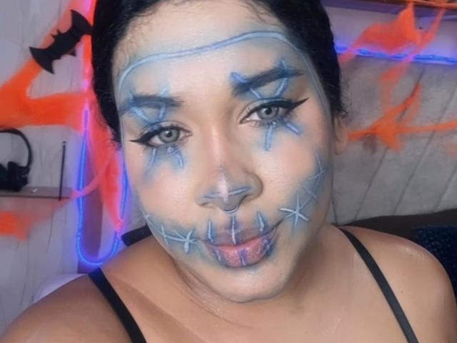 LianaAndAsaff has blue face paint with cross stitches on her forehead, nose, and cheeks, and a stitched smile, wearing black straps, looking at the camera.