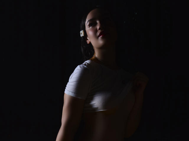 MariaLoft, con cabello oscuro, top corto blanco, pechos pequeños, piel clara, fondo sombrío, mirando hacia arriba, sosteniendo una camisa, iluminación oscura, labios ligeramente entreabiertos, un pasador blanco en la oreja izquierda..