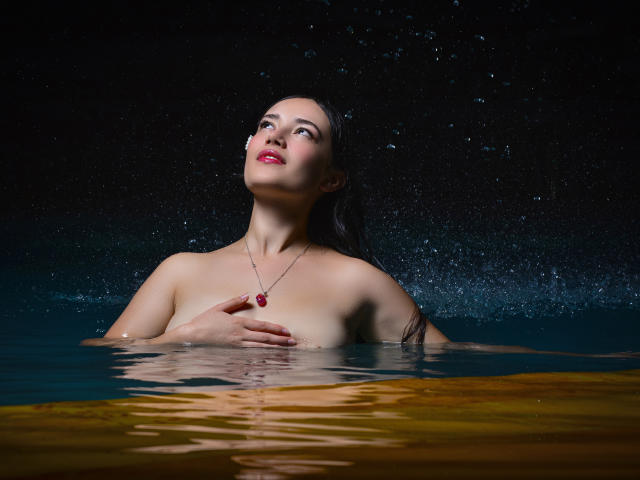 MariaLoft is in a dark, wet pool, covering her tits with one hand, looking up with red lips, wearing a red gem necklace, water droplets around her.