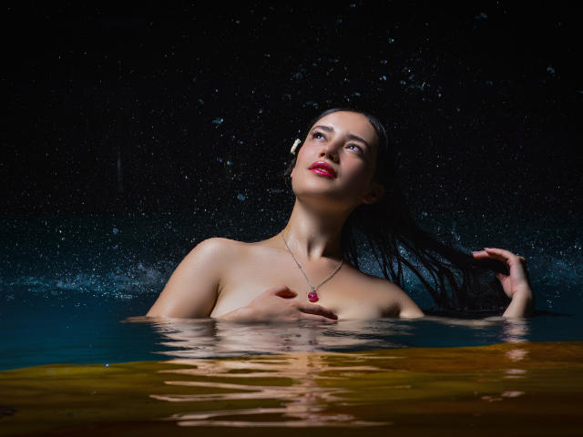 MariaLoft with wet black hair, red lipstick, and necklace, partially submerged in water, hand on chest, looking up, dark background, water droplets, bare breasts, dark skin.