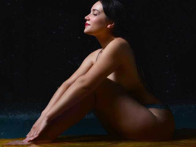 MariaLoft, topless, sitting on yellow surface, knees up, wet skin, dark hair, red lipstick, small grey panties, hands on knees, dark background, water droplets on skin.