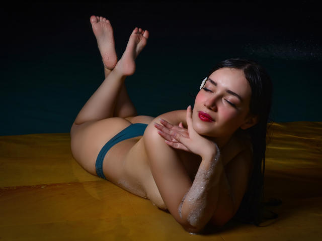 MariaLoft acostada sobre una superficie amarilla, con las piernas levantadas, vistiendo ropa interior turquesa, con las manos en la mejilla, rostro relajado, piel brillante por el agua, lápiz labial rojo, cabello oscuro, fondo negro..
