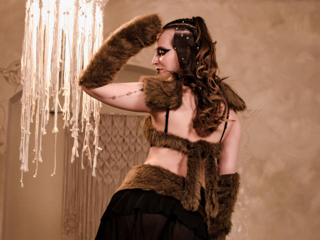 AmbeerWood with brown fur arm sleeves, backless black dress, and brown fur waist, standing under a chandelier, long curly brown hair, black headband, tattoo on right arm.