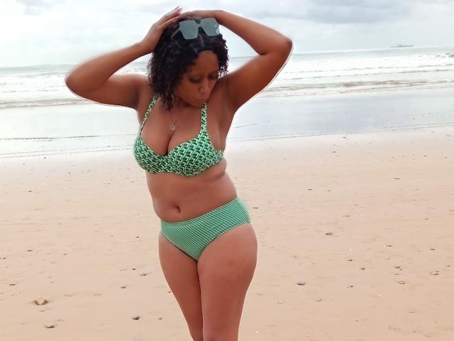 LisaLena with curly black hair, wearing a green patterned bikini, standing on a beach, hands on head, sunglasses on top, tan skin, curvy with medium tits and a soft ass.