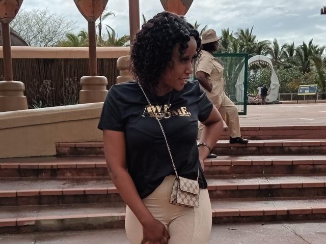 LisaLena has curly black hair, dark shirt showing "MADE" in gold, and white pants. She's holding a quilted purse, standing on brick steps with a man in a tan outfit.